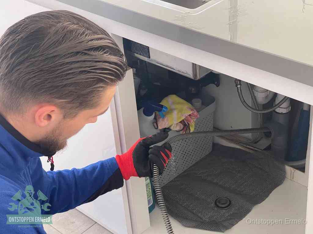 Een monteur voert een flexibele ontstoppingsveer in onder het keukenkastje, langs de sifon, om de gootsteen te ontstoppen in Ermelo