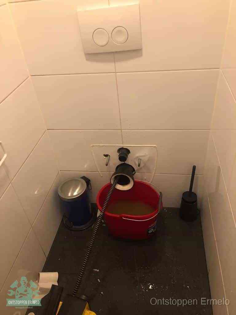 Een ontstopte wand-wc met verwijderde toiletpot waar een ontstoppingsveer in de wanduitgang verdwijnt, het achterhalen waar de verstopping zit in Ermelo
