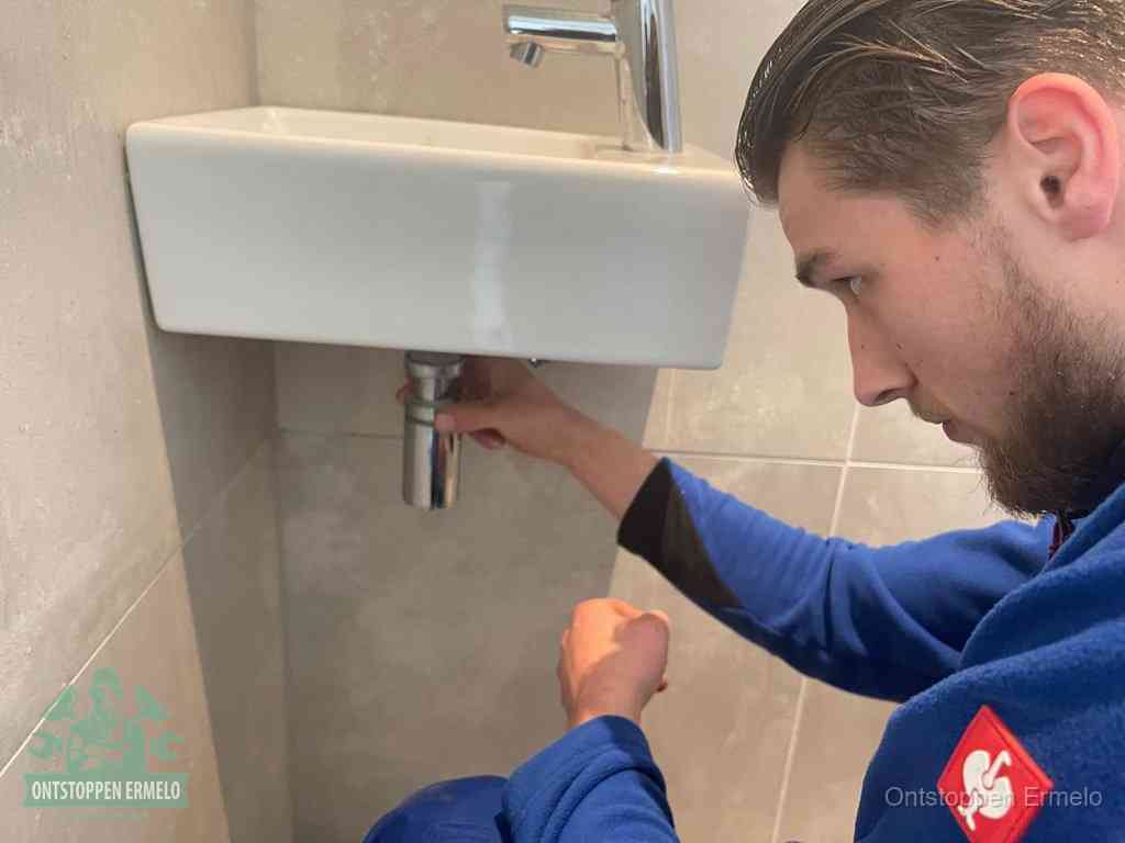 Een monteur draait met de hand de zwanenhalssifon onder een fonteinwasbak in de toiletruimte los om de bron van rioollucht in Ermelo te vinden
