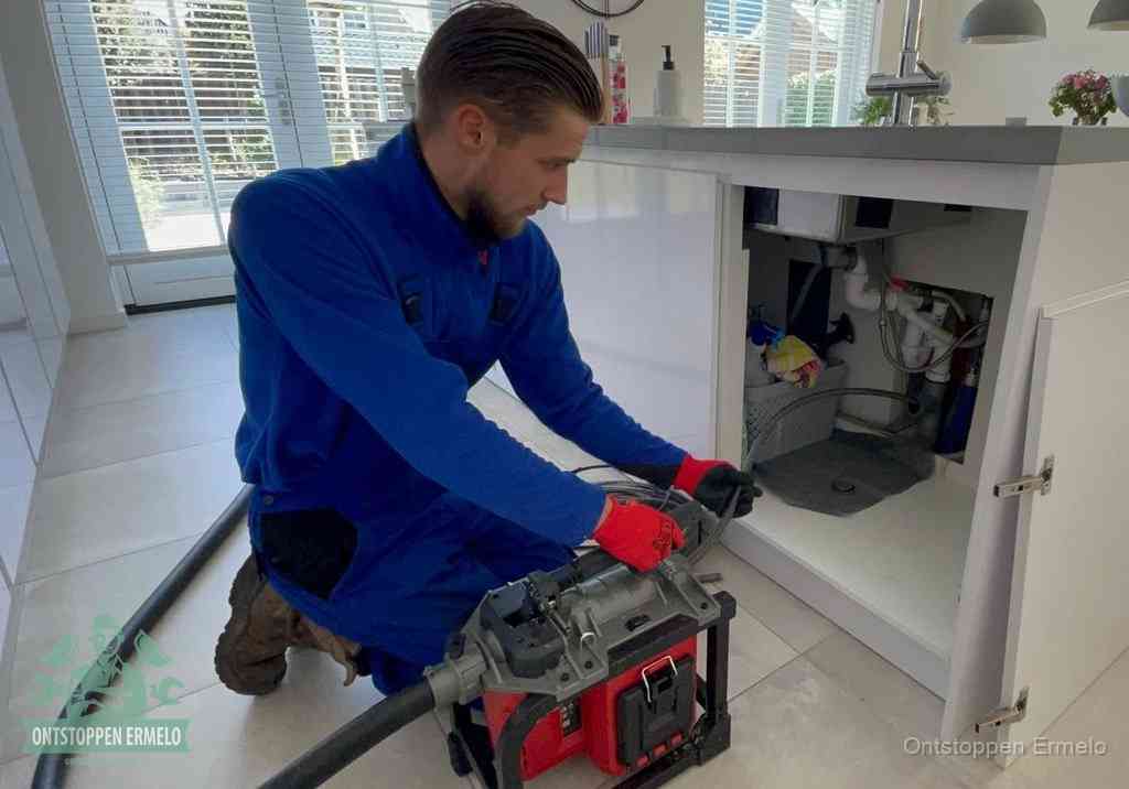 Een monteur voert een professionele elektrische ontstoppingsmachine in de afvoer onder de spoelbak tijdens het gootsteen ontstoppen in Ermelo